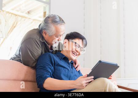 Asiatischer älterer Vater, der sich hinter seinem Sohn mittleren Alters mit einer Brille in der Hand eines Tablets auf der Couch zu Hause liegend Stockfoto