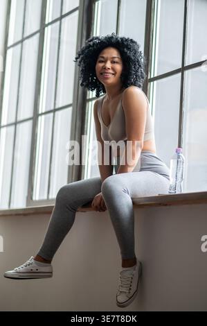 Schwarze Frau mit Locken, die auf der Fensterbank neben der Wasserflasche sitzt Stockfoto