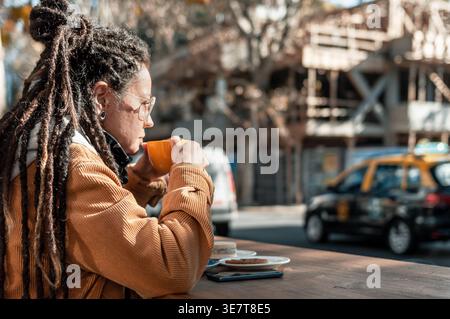 Hispanische Frau mit Dreadlocks, die eine Brille trägt und Kaffee trinkt, sitzt am Tisch im Café im Freien Stockfoto