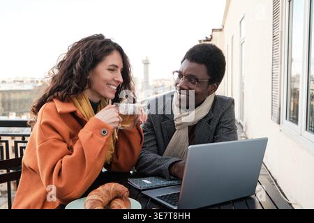 Schwarzer Mann und weiße Frau, die Tee trinken, sitzen am Tisch mit Laptop im Café Stockfoto