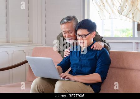 Asiatischer Seniorenvater, der hinter ihm steht und auf den Laptop blickt, mit einem Mann mittleren Alters, der zu Hause auf der Couch sitzt Stockfoto