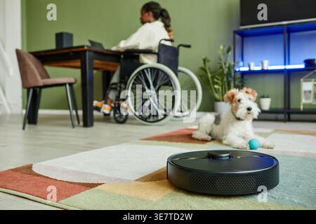 Behinderte Schwarze Frau im Rollstuhl sitzt am Tisch, arbeitet an einem Laptop in der Nähe ihres Haustierhundes, der zu Hause auf Teppich sitzt Stockfoto
