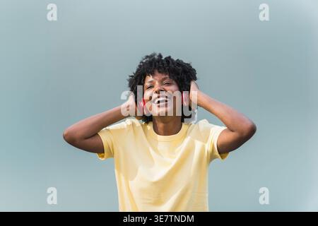 Schwarze Frau in gelbem Hemd, die Musik auf kabellosen Kopfhörern hört, während sie im Studio vor verschwommenem Hintergrund steht Stockfoto