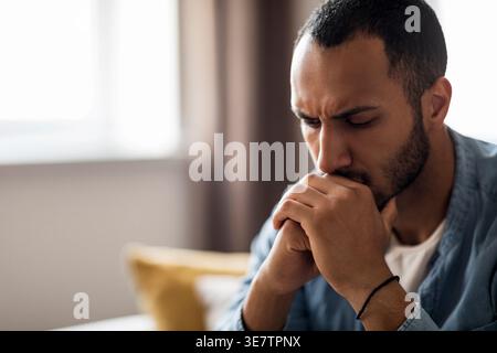 Trauriger Schwarzer Mann, der den Mund auf gefesselte Hände lehnt, sitzt auf Sofa im Wohnzimmer zu Hause Stockfoto