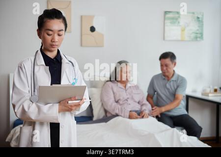 Asiatische Ärztin, die die medizinischen Aufzeichnungen des Patienten füllt, nachdem sie die ältere Frau mit ihrem Mann überprüft hat, der auf dem Bett des Krankenhauszimmers sitzt Stockfoto