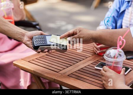 Hand eines asiatischen männlichen Kellners, der kontaktlose Zahlungen an eine mittelalte Schwarze Frau, die mit einer Debitkarte bezahlt, am Tisch sitzt mit weiblichen Freunden im Outd... Stockfoto