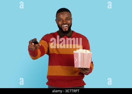 Fröhlicher Schwarzer, der Popcorn-Eimer mit Fernbedienung hält, während er im Studio vor blauem Hintergrund steht Stockfoto