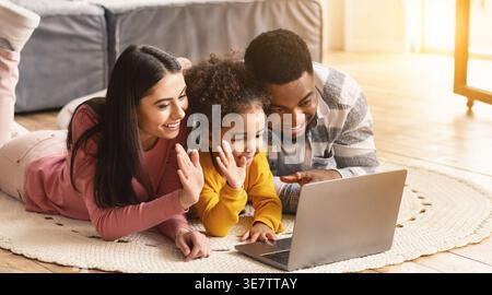 Lächelnde Schwarze und kaukasische Eltern mit ihrer Tochter, die die Hände winkt und Videoanrufe auf dem Laptop macht, während sie zu Hause auf dem Teppich liegen Stockfoto