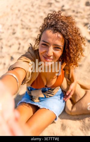 Frau aus dem Nahen Osten mit lockigen Haaren in Sommerkleidung, die tagsüber Selfie macht, während sie am Strand sitzt Stockfoto