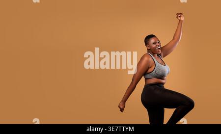 Fröhliche schwarze Frau mit kurzen Haaren, die Sportswear trägt, die geballte Faust hochhebt, während sie im Studio vor braunem Hintergrund tanzt Stockfoto
