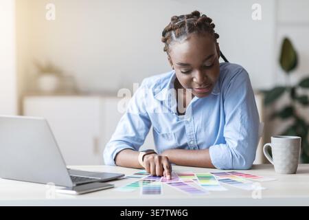 Schwarze Geschäftsfrau mit Zöpfen, die sich die Farbpalette anschauen, während sie im Home Office am Tisch mit Laptop sitzt Stockfoto