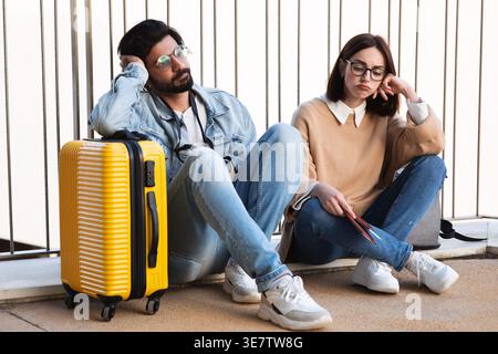 Asiatische und kaukasische Paare mit Brille und Kopf auf den Händen, die wegschauen und Reisedokumente halten, während sie zusammen auf Beton sitzen... Stockfoto