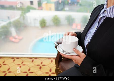 Zugeschnittenes Bild einer chinesischen Rezeptionistin mit Kaffeetasse, die in der Hotellobby auf einem Stuhl sitzt Stockfoto