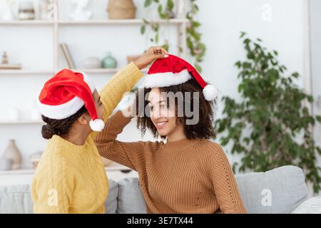 Vorbereitung auf Neujahr und Weihnachten und Party zu Hause. Fröhliche afroamerikanerin in Pullover zieht ihrem Freund Weihnachtsmütze an und ist bereit für ... Stockfoto