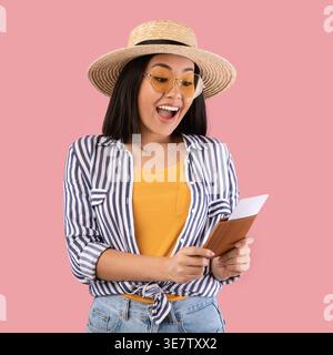 Fröhliche asiatische Frau mit Strohhut und Sonnenbrille, die den Pass mit Flugtickets hält und ansieht, während sie ... vor rosa Hintergrund stand Stockfoto