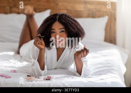 Lächelnde Schwarze Frau mit lockigem Haar, die einen Bademantel trägt, nagellack aufträgt, um Maniküre zu machen, während sie auf dem Bauch auf dem Bett zu Hause im Schlafzimmer liegt Stockfoto