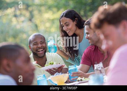 Lächelnder schwarzer Mann mit Teller und Essen am Tisch neben einer spanischen Freundin mit einem Glas Wasser am Tisch neben männlichen Kollegen ... Stockfoto