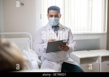 Nahöstlicher Arzt mit Gesichtsmaske, der im Krankenhaus liegender Patient befragt Stockfoto