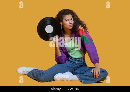 Afroamerikanerin, die Vinyl-Schallplatte in der Nähe des Halses hält und drinnen auf dem Boden sitzt Stockfoto