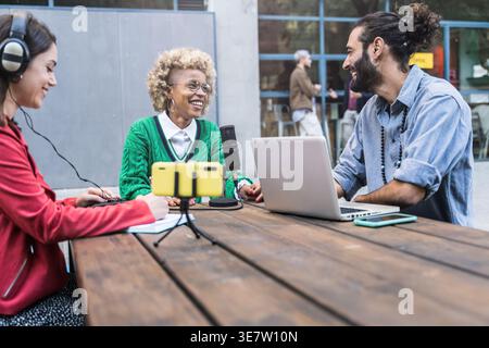 Lächelnde Schwarze Frau mit kaukasischen Kollegen, die tagsüber am Tisch sitzen und ein Interview für Online-Podcast in einem Restaurant im Freien aufzeichnen Stockfoto