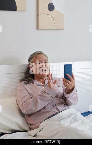 Asiatische ältere Frau mit Smartphone-Videoanruf relativ winkende Hand, während sie auf dem Bett im Krankenhauszimmer sitzt Stockfoto