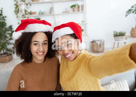 Lächelnd verschiedene weibliche Freunde mit Weihnachtsmützen und Pullover, die zu Hause auf dem Sofa sitzen Stockfoto