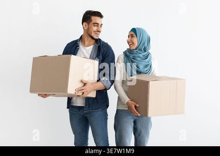 Mann aus dem Nahen Osten und muslimische Frau im Hijab, die Papierkisten tragen und sich gegenseitig in ihrem neuen Haus ansehen Stockfoto