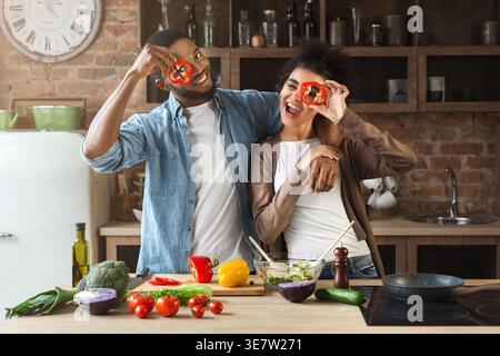 Lächelndes schwarzes Paar, das in Scheiben geschnittene Paprika hält und Salat zubereitete, während er am Tisch in der Küche zu Hause steht Stockfoto