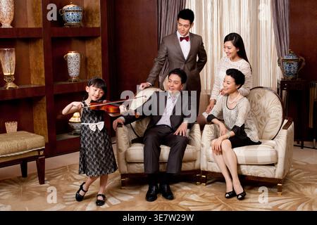 Lächelndes chinesisches Mädchen, das vor Familienmitgliedern auf dem Sofa steht und Geige spielt und sie zuhause beobachtet Stockfoto