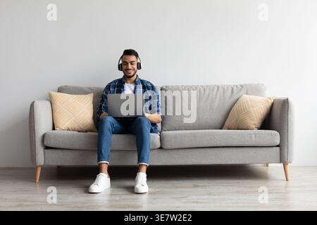 Lächelnder Mann aus dem Nahen Osten, der Kopfhörer trägt, während er am Laptop arbeitet und zu Hause auf dem Sofa sitzt Stockfoto