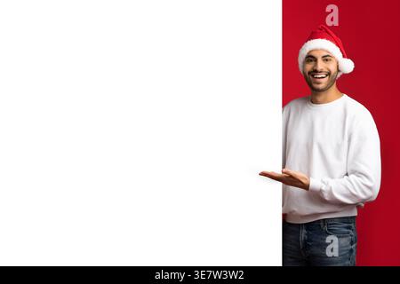 Lächelnder Mann aus dem Nahen Osten mit Weihnachtsmann-Hut, der Whiteboard zeigt, während er vor rotem Hintergrund im Studio steht Stockfoto