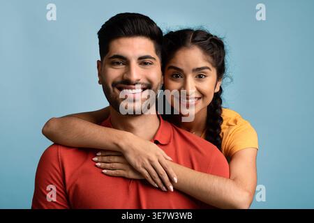 Ein lächelnder Mann aus dem Nahen Osten mit seiner Freundin, die ihn von hinten umschließt, während er im Studio vor blauem Hintergrund steht Stockfoto