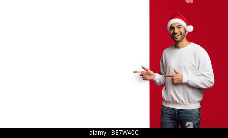Ein lächelnder Mann aus dem Nahen Osten trägt einen Weihnachtsmann und zeigt mit den Fingern auf dem Whiteboard, während er im Studio vor rotem Hintergrund steht Stockfoto
