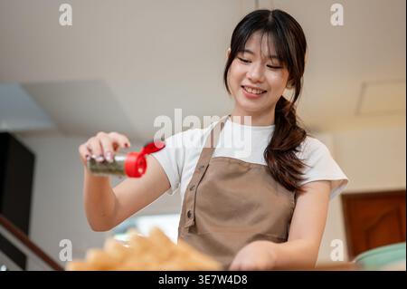 Thailändische Frau mit Schürze, die Pfeffer auf ihr Essen streut, während sie zu Hause in der Küche steht Stockfoto