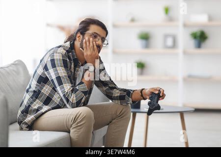 Arabischer Mann mit Brille, der den Spielcontroller mit dem Kinn an der Hand hält, während er zu Hause auf dem Sofa sitzt Stockfoto