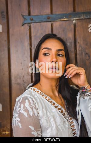 Ernsthafte hispanische Frau, die das Gesicht berührt und wegschaut, während sie an einer Holzwand steht Stockfoto