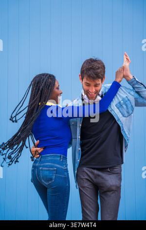 Lächelnde Schwarze und kaukasische Paare mit Kopfhörern um den Hals, die Hände halten und Salsa vor blauem Hintergrund im Studio tanzen Stockfoto