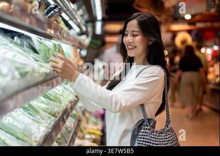 Thailändische Frau mit Schultertasche, die Gemüse aus dem Regal wählt, während sie im Supermarkt steht Stockfoto