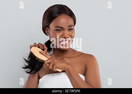 Verärgerte Schwarze Frau, die in Handtuch gewickelt ist, das verworrene Haare kämmt, die vor grauem Hintergrund im Studio stehen Stockfoto