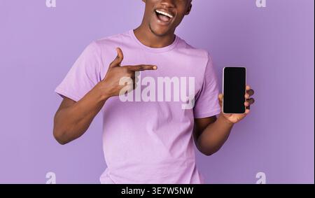 Schwarzer Mann, der mit dem Finger auf das Smartphone zeigt, während er im Studio vor violettem Hintergrund steht Stockfoto