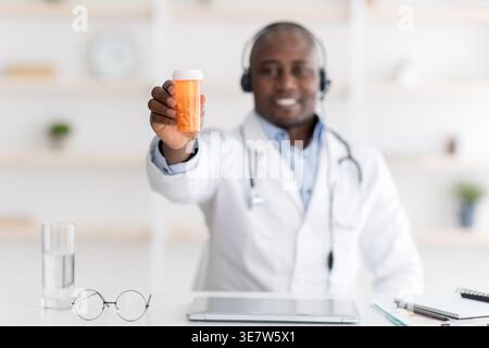 Schwarzer männlicher Arzt, der ein Headset trägt, das eine Arztdose zeigt, während er in der Klinik mit Tablet und Brille am Schreibtisch sitzt Stockfoto