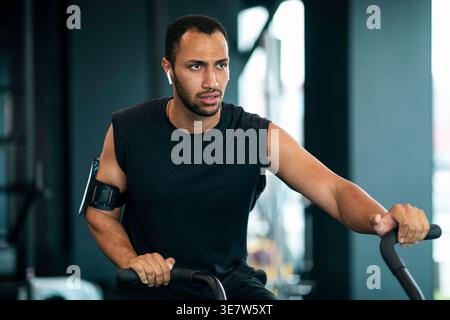 Schwarzer Mann mit Bart, der schnurlose Ohrhörer und Armband trägt und wegschaut, Training mit einem Crosstrainer im Fitnessstudio Stockfoto