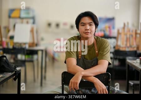 Lächelnder asiatischer männlicher Künstler auf Schürze sitzend und auf einem Stuhl in der Handwerkstatt Stockfoto