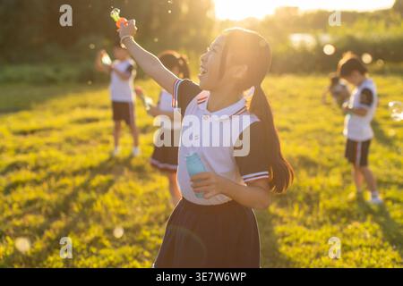 Fröhliche chinesische Schülerin in Schuluniform, die an sonnigem Tag neben Klassenkameraden am Seeufer eine Flasche spielt Stockfoto
