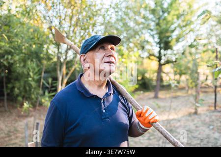 Lächelnder hispanischer männlicher Bauer in blauem Hemd und Mütze mit Handschuhen, der tagsüber auf dem Bauernhof steht Stockfoto