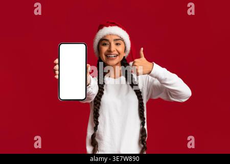 Cool App Fröhliche Junge Arabische Frau In Santa Hat Zeigt Leeres Smartphone Mit Weißem Bildschirm Und Daumen Nach Oben, Joyful Middle Eastern Female Recom Stockfoto