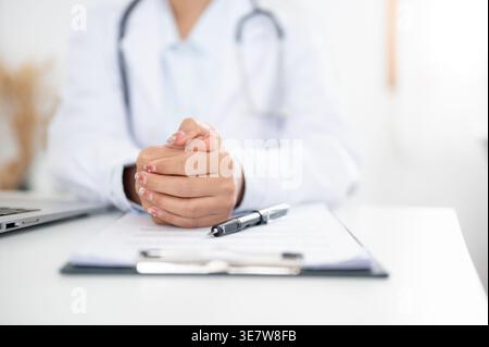 Thailändische Ärztin mit gefesselten Händen am Tisch mit Laptop, Klemmbrett und Stift in der Klinik Stockfoto