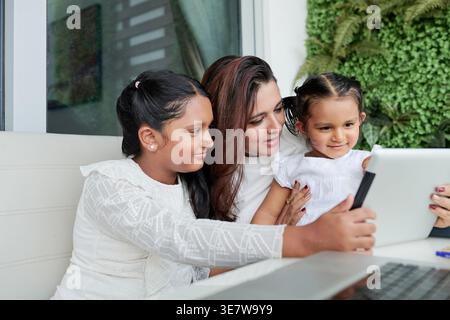 Indische Mutter und Töchter, die zusammen ein Tablet benutzen, sitzen am Tisch im Hinterhof Stockfoto
