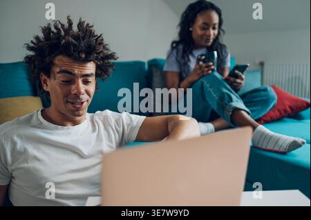 Mann mit gemischter Rasse, der an Laptop in der Nähe von Schwarzer Freundin arbeitet, die Kaffeetasse hält, während sie das Smartphone benutzt, das auf dem Sofa im Wohnzimmer zu Hause sitzt Stockfoto