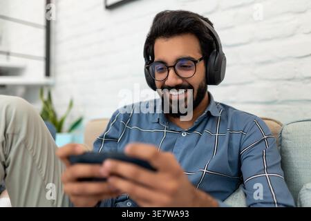 Lächelnder bärtiger indischer Mann mit Brille und Kopfhörern, der Online-Spiel spielt und auf Sofa im Wohnzimmer zu Hause sitzt Stockfoto